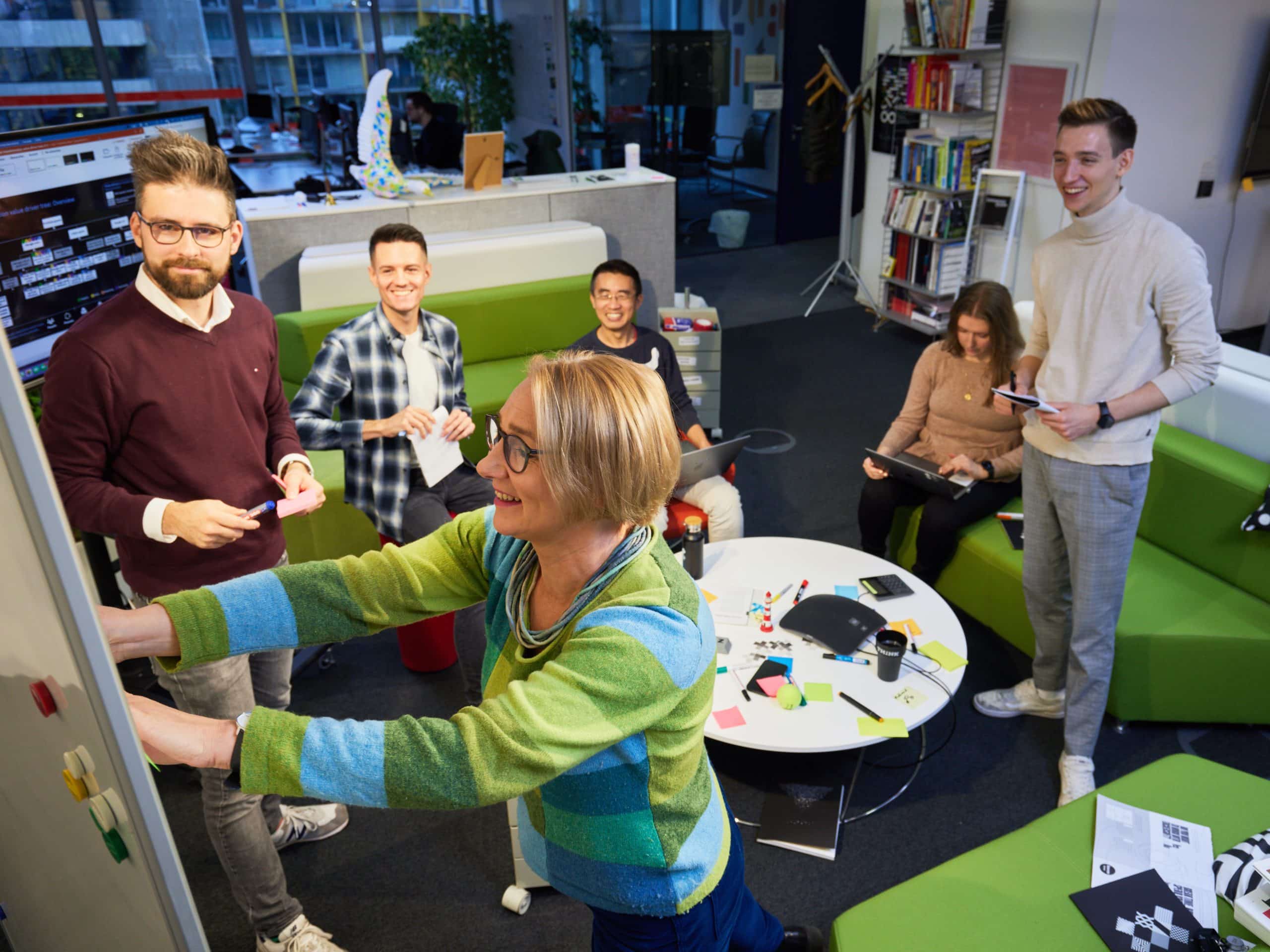 Vier Menschen arbeiten in der Gruppe. Drei sitzen und eine Frau steht an der Tafel und klebt ein Post-It ran.